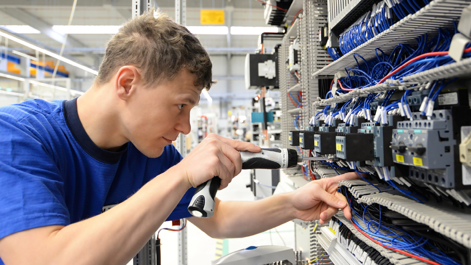 A man wearing a bright blue shirt uses a tool to fix a component on an electrical panel.