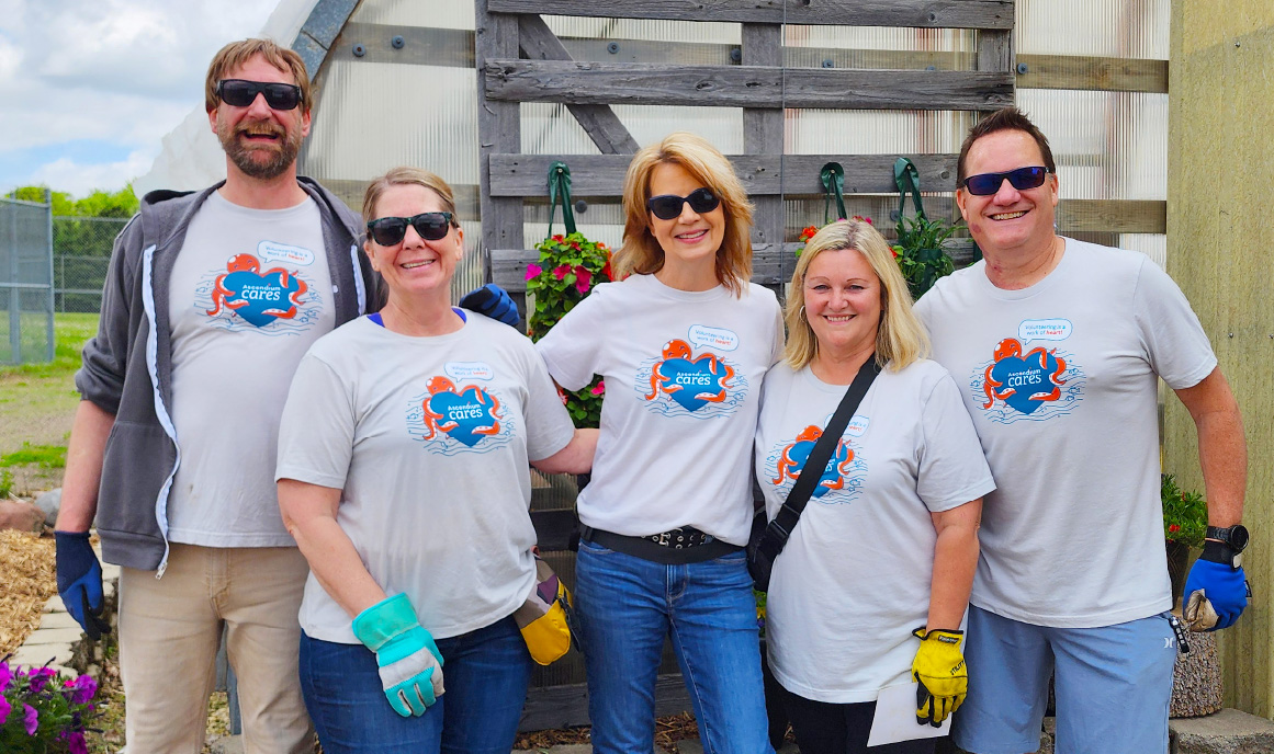 A smiling group of five Ascendium volunteers wearing work gloves and Ascendium Cares t-shirts stand in front of hanging flowers and a greenhouse